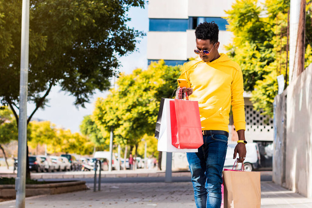 Sarı eşofman altı, kot pantolon ve güneş gözlüğü giymiş genç Afrikalı bir adam elinde alışveriş torbalarıyla sokakta yürürken cep telefonuna bakıyor.. - Fotoğraf, Görsel