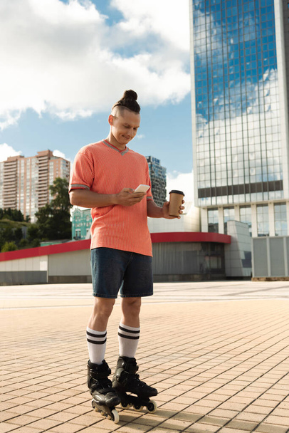 Χαμογελώντας πατινέρ πατινάζ χρησιμοποιώντας smartphone και κρατώντας καφέ για να πάει στο αστικό δρόμο  - Φωτογραφία, εικόνα