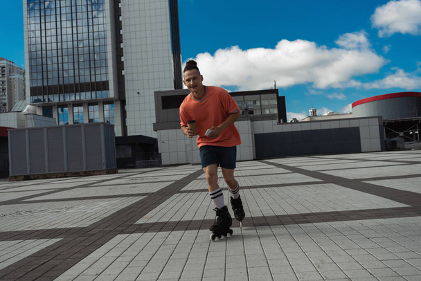 Cheerful man in roller skates holding smartphone and coffee to go on city street  - Photo, Image