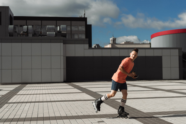 Молодой человек в роликовых коньках держит кофе, чтобы пойти и смартфон во время езды по городской улице  - Фото, изображение