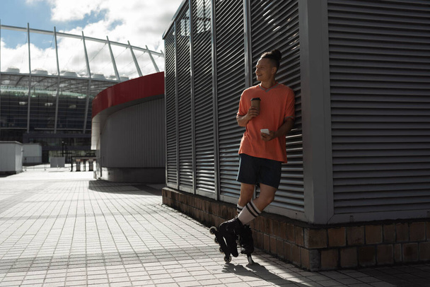 Vreugdevolle rolschaatser met smartphone en koffie te gaan in de buurt van gebouw op stedelijke straat  - Foto, afbeelding
