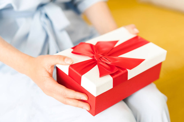 Mujer sentada y sosteniendo delante de su hermosa caja de regalos con lazo rojo en las manos. Caja regalo festiva roja y blanca en las manos de la mujer. Sin cara. Enfoque selectivo - Foto, imagen