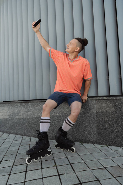 Vue latérale de l'homme souriant en patins à roulettes prenant selfie sur smartphone près du bâtiment à l'extérieur  - Photo, image