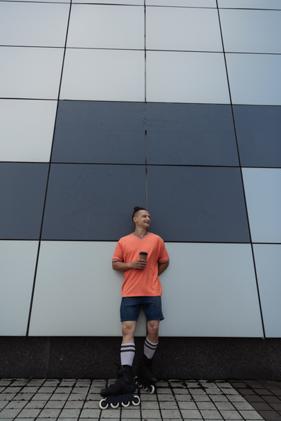 Wide angle view of smiling roller skater holding coffee to go near building outdoors  - Photo, Image