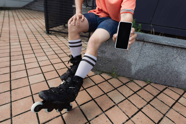 Rajattu näkymä miehen rullat ja rento vaatteita osoittaa matkapuhelin tyhjällä näytöllä ulkona  - Valokuva, kuva