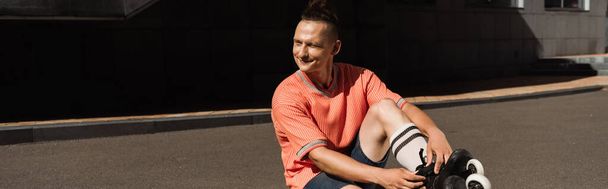 Hombre sonriente en camiseta y patines sentado en asfalto al aire libre, pancarta  - Foto, Imagen