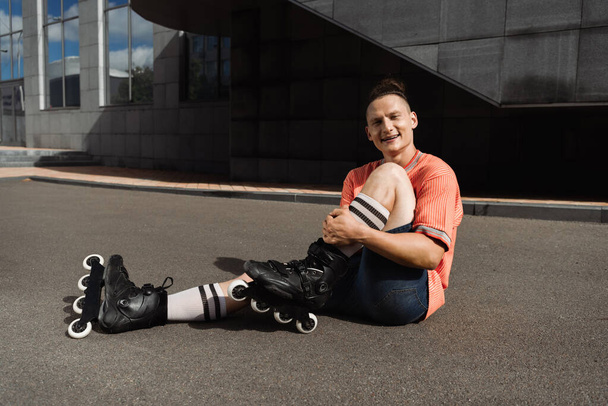 vrolijke en stijlvolle rolschaatser zittend op asfalt en kijkend naar camera - Foto, afbeelding