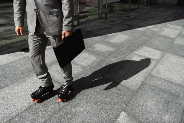 vue recadrée de l'homme d'affaires en patins à roulettes et costume gris tenant une mallette noire dans la rue en ville - Photo, image