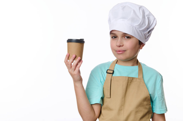 Pohledný středovýchodní dospívající chlapec v kuchařské čepici a béžové zástěře, prodávající odnášející horký nápoj kávový čaj v eko papírovém kelímku jít, roztomile se usmívá při pohledu do kamery, izolované přes bílé pozadí - Fotografie, Obrázek