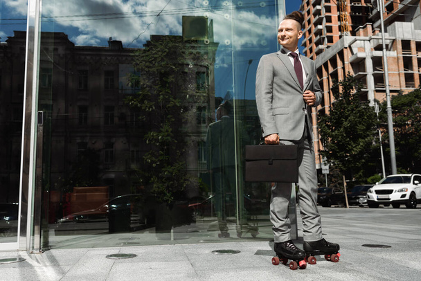 улыбающийся бизнесмен в роликовых коньках, держащий черный портфель и смотрящий в сторону на городской улице - Фото, изображение