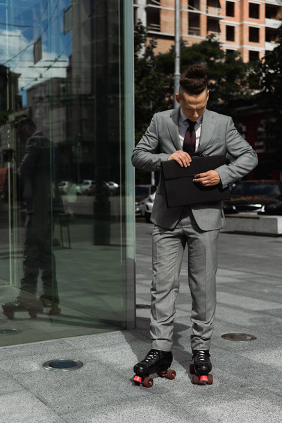 volle Länge des stilvollen Mannes in formaler Kleidung und Rollschuhen, der die Aktentasche auf der Stadtstraße schließt - Foto, Bild