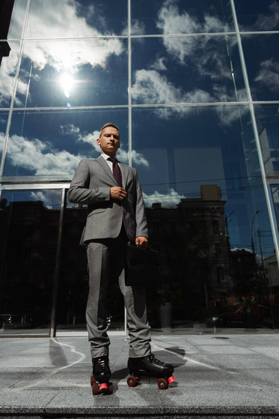 low angle view of confident businessman in grey suit and roller skates near contemporary building with glass facade - Фото, зображення