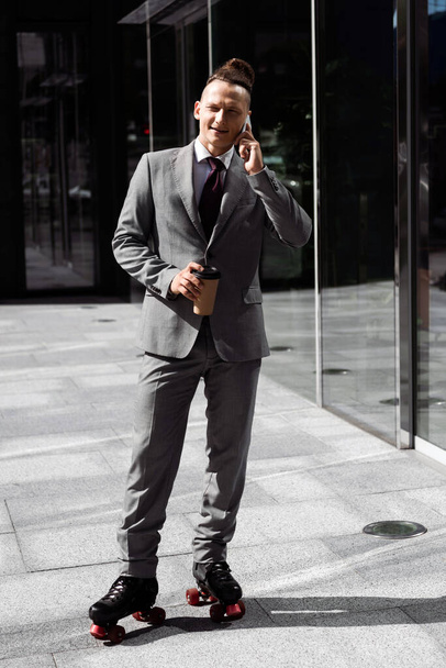 full length of businessman in suit and roller skates holding coffee to go and talking on smartphone outdoors - Photo, Image
