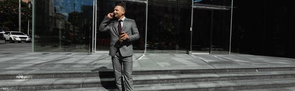 elegante hombre de negocios en traje gris celebración de bebidas para llevar y hablar en el teléfono celular en la calle de la ciudad, pancarta - Foto, Imagen
