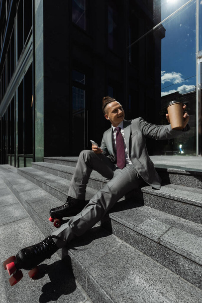 Lächelnder Geschäftsmann in Anzug und Rollschuhen sitzt mit Smartphone und Kaffee to go auf der Treppe und schaut draußen weg - Foto, Bild