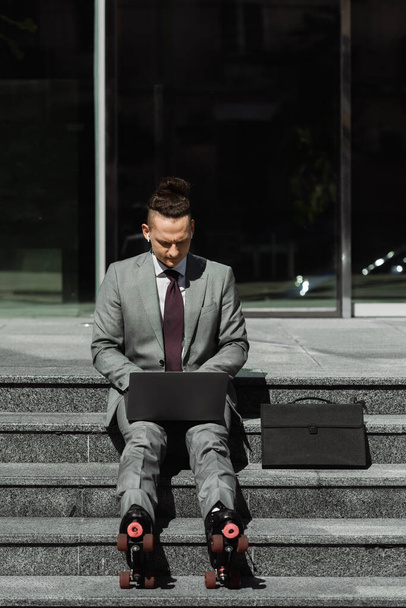 homem de negócios elegante no desgaste formal e patins digitando no laptop enquanto sentado em escadas perto da pasta - Foto, Imagem