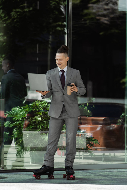 smiling man in grey suit and roller skates holding coffee to go and looking at laptop near green plant behind glass facade - Fotografie, Obrázek