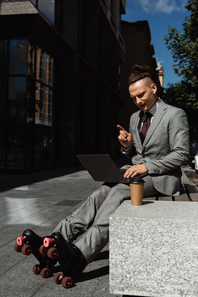 young man in roller skates and business suit sitting with laptop near coffee to go and pointing with finger - Photo, Image