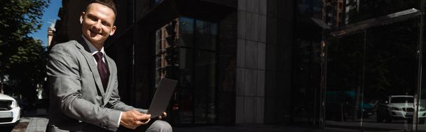 boldog üzletember szürke öltönyben ül laptoppal és nézi a kamerát a városi utcán, banner - Fotó, kép