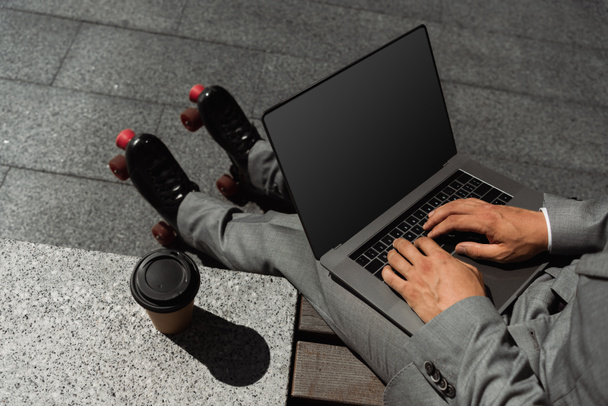 top view of cropped businessman in roller skates typing on laptop with blank screen near coffee to go outdoors - Photo, Image