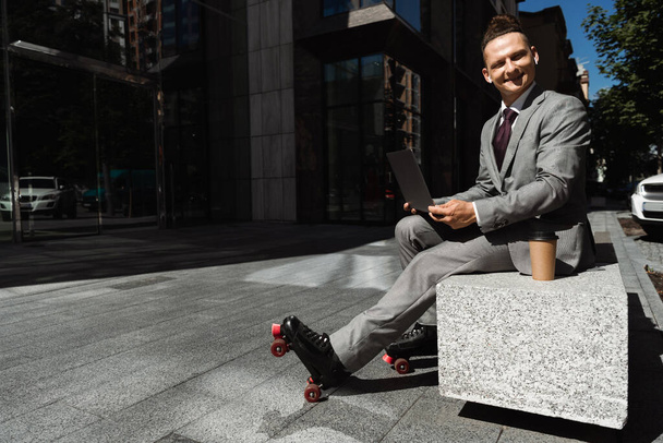 smiling man in roller skates and formal wear looking at camera while sitting with laptop on street - Photo, Image