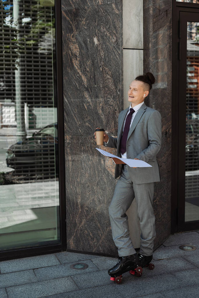 homme d'affaires souriant en patins à roulettes debout avec des documents et du café pour aller près du bâtiment dans la rue - Photo, image