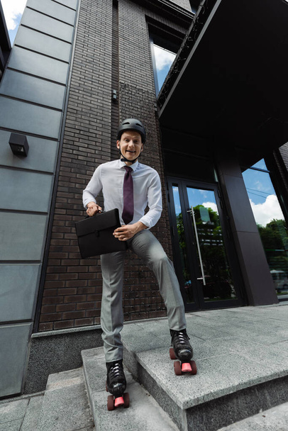cheerful roller skater in formal wear and helmet holding briefcase and smiling at camera near modern building - Photo, Image