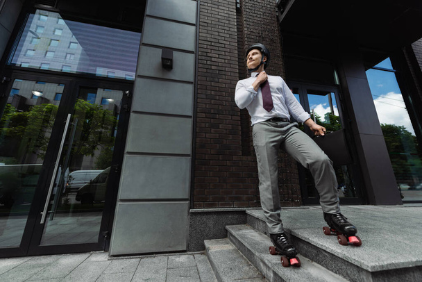 longitud completa de hombre de negocios en patines de ruedas y corbata de fijación de desgaste formal y mirando hacia otro lado cerca de edificio contemporáneo - Foto, imagen