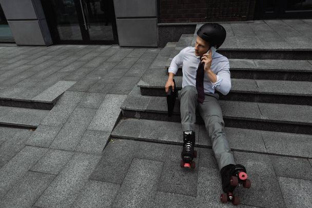 vista ad alto angolo di uomo d'affari in casco e pattini a rotelle parlando su smartphone mentre seduto sulle scale all'aperto - Foto, immagini