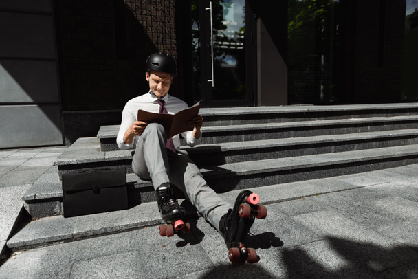 jeune homme d'affaires en patins à roulettes et casque regardant le document tout en étant assis sur les escaliers à l'extérieur - Photo, image