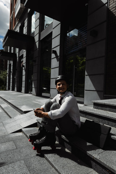 homme d'affaires heureux en patins à roulettes et casque tenant smartphone et souriant à la caméra tout en étant assis sur les escaliers près du bâtiment  - Photo, image