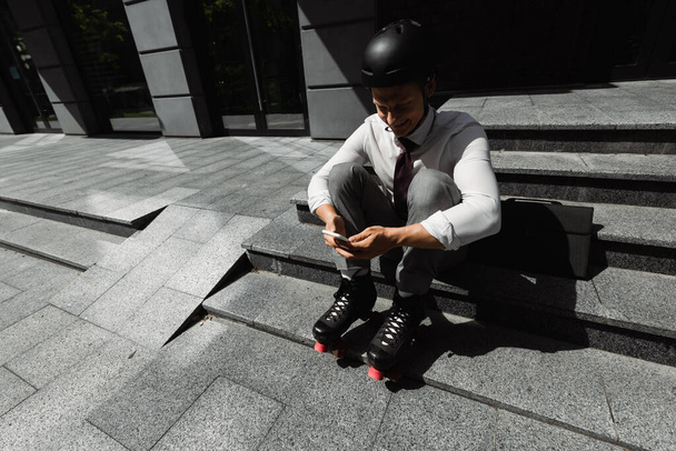 vue grand angle de l'homme d'affaires en patins à roulettes assis sur les escaliers et la messagerie sur smartphone - Photo, image