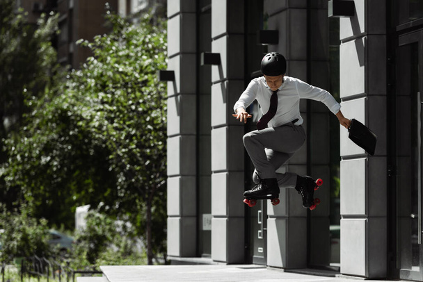 full length of businessman in roller skates and helmet jumping while doing trick on street near building - Valokuva, kuva