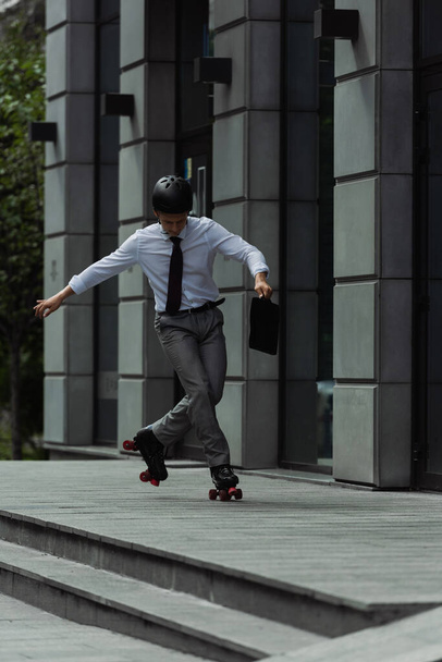 full length of man in formal wear and roller skates doing trick near modern building on city street - Foto, afbeelding