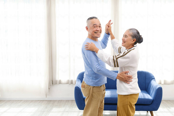 Ρομαντικό και χαρούμενο ζευγάρι ηλικιωμένων από την Ασία απολαμβάνει να χορεύει και να κρατάει το χέρι της μουσικής μαζί με χαμόγελα και ευτυχία στο σαλόνι. Ανώτερη δραστηριότητα συνταξιοδότησης στο σπίτι. - Φωτογραφία, εικόνα