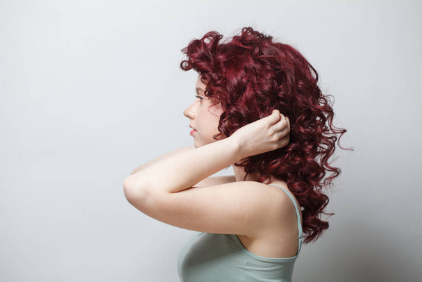 Mujer atractiva tocando su pelo rizado rojo contra fondo blanco de la pared - Foto, Imagen