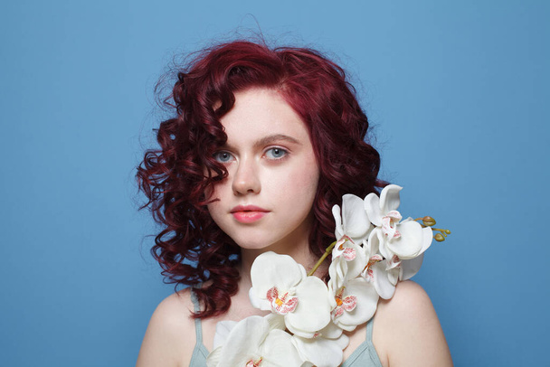 Приваблива жінка з червоним кучерявим волоссям, що тримає білу орхідею на фоні синьої стіни студії
 - Фото, зображення