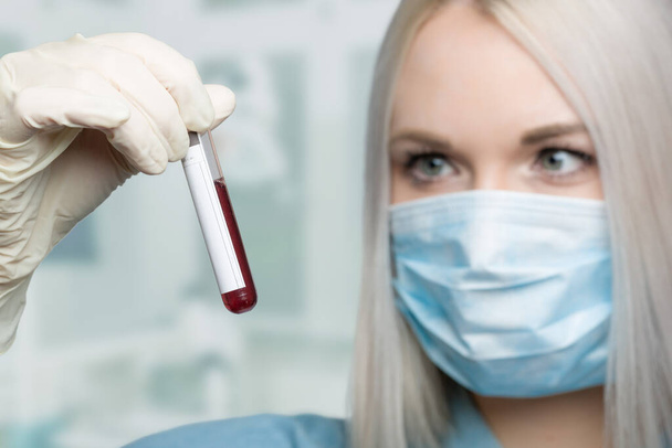 医療用フェイスマスクと医療用手袋を身に着けている若い女性は研究室で血液プローブを扱っています  - 写真・画像