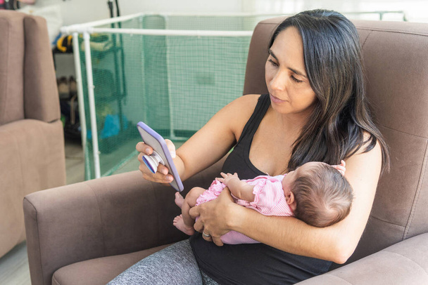 Reife Frau mit ihrem Neugeborenen im Arm, das zu Hause vom Handy aus arbeitet - Foto, Bild