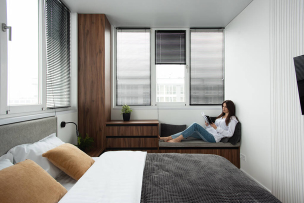 Mujeres lectura libro en un dormitorio con cama y armario de madera en apartamento moderno. persianas en las ventanas - Foto, imagen