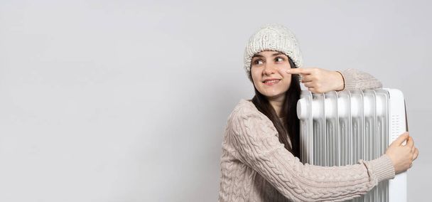A woman in a hat hugs an oil heater and points her finger to the side in place for text - Foto, Imagem