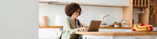 African american young woman with curly afro hairstyle and eyeglasses using laptop at home - Фото, изображение