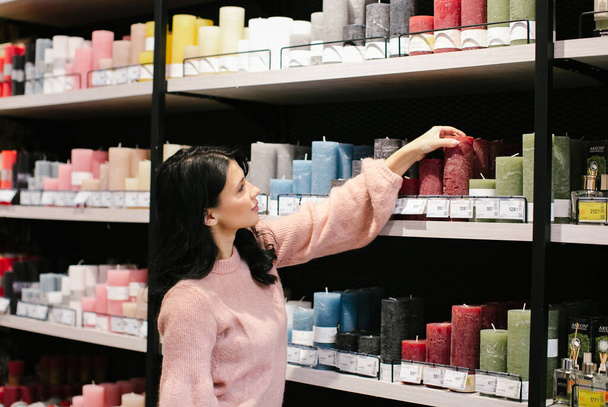A young brunette woman in a pink sweater buys a decorative candle in a store. - Fotoğraf, Görsel