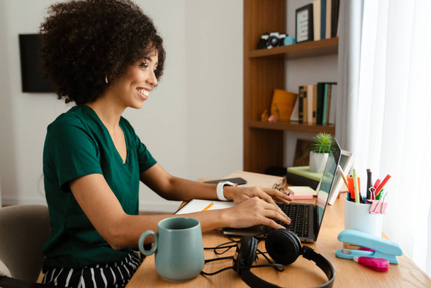African american young woman with curly afro hairstyle sitting at desk and using laptop at home - Photo, image