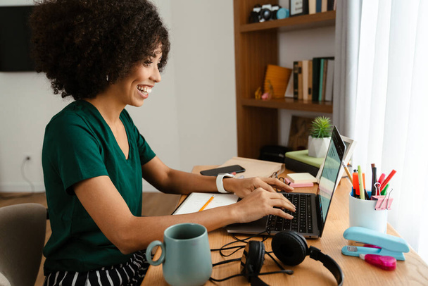 African american young woman with curly afro hairstyle sitting at desk and using laptop at home - 写真・画像