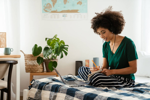 African american young woman taking notes while sitting in bed and using laptop at home - Foto, afbeelding