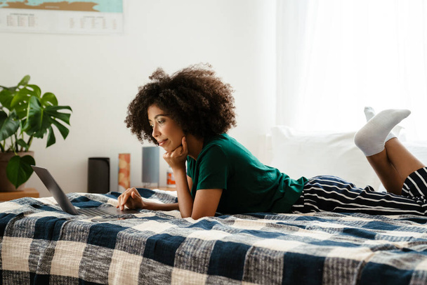 African american young woman with curly hairstyle lying in bed and using laptop at home - Fotografie, Obrázek