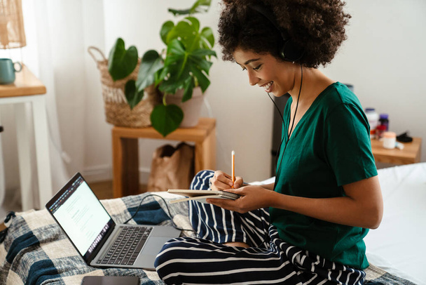 African american young woman taking notes while sitting in bed and using laptop at home - Valokuva, kuva