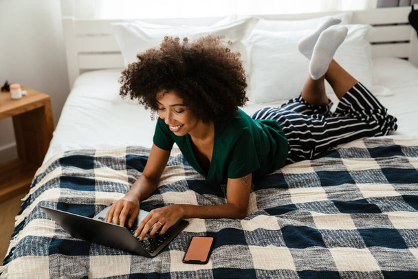 African american young woman with curly hairstyle lying in bed and using laptop at home - Valokuva, kuva