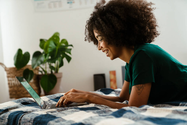 African american young woman with curly hairstyle lying in bed and using laptop at home - Φωτογραφία, εικόνα